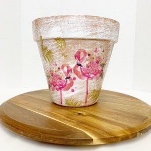 May include: A terracotta planter with a white distressed finish and a pink flamingo design. The planter is decorated with gold leaf accents and pink flowers.