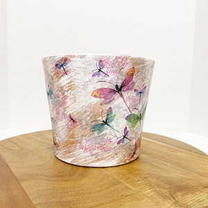 May include: White ceramic planter with a floral pattern featuring pink, purple, and blue dragonflies. The planter is sitting on a wooden surface.