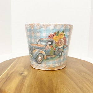 May include: A blue and white ceramic planter with a vintage truck design. The truck is carrying a bouquet of flowers in shades of yellow, orange, and pink. The planter has a checkered pattern and a distressed finish.