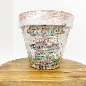 May include: A white terracotta planter with a beach theme design. The planter has a distressed white finish and features a sign that says "to the beach" with arrows pointing to other destinations, including "South Island", "Boat Rentals", "Beach Cottage", "Cala Beach", and "Habana".