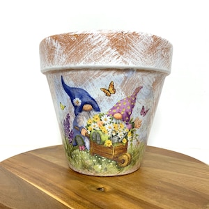 May include: A decorative terracotta flower pot featuring a whimsical garden scene. Two gnomes are pushing a wheelbarrow filled with yellow and white flowers. Butterflies and lavender accents adorn the light blue background. The rim has a distressed, white-washed appearance.