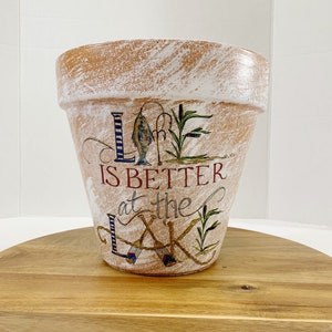 May include: A white terracotta flower pot with a distressed finish. The pot is decorated with a hand-painted design of a fish, fishing poles, and the text "Life is better at the lake".