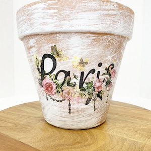 May include: A white terracotta flower pot with a distressed finish. The pot is decorated with a floral design, butterflies, and the word "Paris" in black lettering.