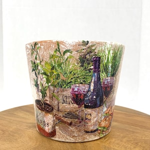 May include: A white ceramic planter with a distressed finish features a colorful illustration of wine bottles, glasses, herbs, and a corkscrew. The planter has a rustic, vintage aesthetic.