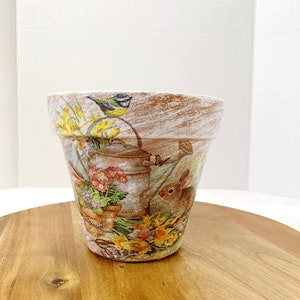 May include: A white ceramic flower pot with a distressed finish. The pot features a colorful floral design with a bluebird, a rabbit, and a watering can.
