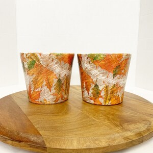 May include: Two small terracotta pots with a fall leaf design. The pots are on a wooden surface.