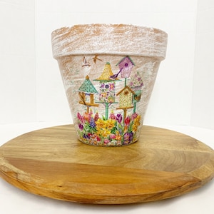 May include: A white terracotta flower pot with a distressed finish, decorated with a colorful design of birdhouses and flowers.