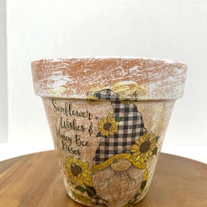 May include: A terracotta flower pot with a distressed white finish. The pot is decorated with a gnome holding sunflowers and the text "Sunflower Wishes & Honey Bee Kisses".