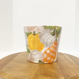 May include: A white ceramic planter with a distressed finish features a colorful autumn-themed design of pumpkins and gourds. The planter is sitting on a wooden surface.