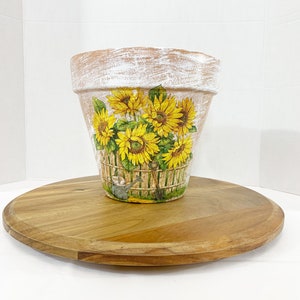 May include: A white terracotta flower pot with a distressed finish, decorated with a colorful image of sunflowers in a basket with a watering can and gardening tools.