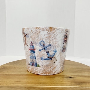 May include: A white ceramic planter with a nautical theme. The planter features a lighthouse, anchor, and a ship's wheel. The planter has a distressed white finish with a brown undercoat.