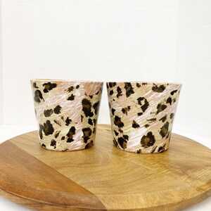 May include: Two ceramic planters with a leopard print design. The planters are on a wooden base.