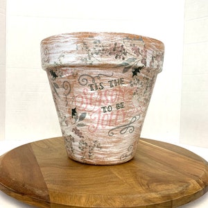 May include: A white terracotta planter with a distressed finish and a floral design. The planter has the text "Tis the Season to be Jolly" written on it.