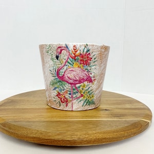 May include: A white ceramic planter with a distressed finish features a pink flamingo and tropical flowers. The planter is sitting on a wooden surface.