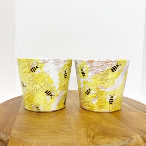 May include: Two yellow and white ceramic flower pots with a painted design of bees and honeycombs. The pots are on a wooden surface.