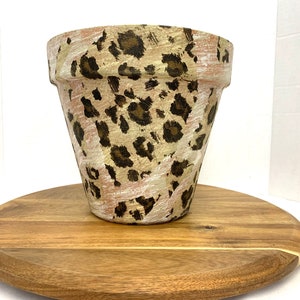 May include: A terracotta planter with a leopard print design in shades of brown, beige, and white. The planter is sitting on a wooden surface.