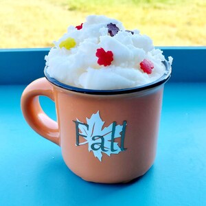 May include: An orange mug with a black rim and handle, filled with white foam and decorated with colorful leaf-shaped sprinkles. The mug has the word "Fall" and a leaf design on the front.