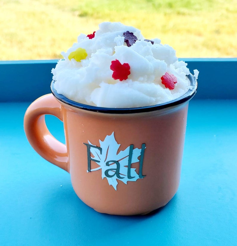 May include: An orange mug with a black rim and handle, filled with white foam and decorated with colorful leaf-shaped sprinkles. The mug has the word "Fall" and a leaf design on the front.