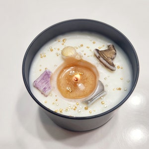 Könnte beinhalten: Eine brennende Kerze in einem schwarzen Behälter. Die Kerze hat eine goldfarbene Mitte und ist von weißem Wachs umgeben. Dekorative Elemente sind ein lila Kristall, ein goldener Hut und goldene Streusel.