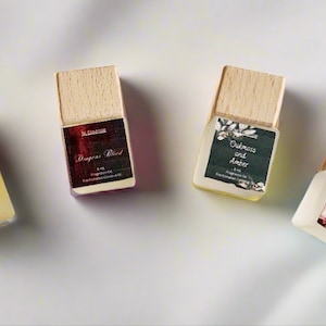 Könnte beinhalten: Vier kleine, rechteckige Duftölflaschen mit Holzverschlüssen sind auf einer weißen Oberfläche angeordnet. Jede Flasche hat ein Etikett mit einem anderen Duftnamen: Citrus Agave, Dragon's Blood, Oakmoss and Amber und Blackberry Sangria. Die Etiketten zeigen botanische Illustrationen.
