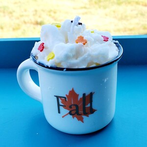 May include: A white enamel mug with a black rim and handle, filled with a white, textured candle. The mug has a "Fall" graphic with a maple leaf. The candle is decorated with colorful leaf-shaped sprinkles.