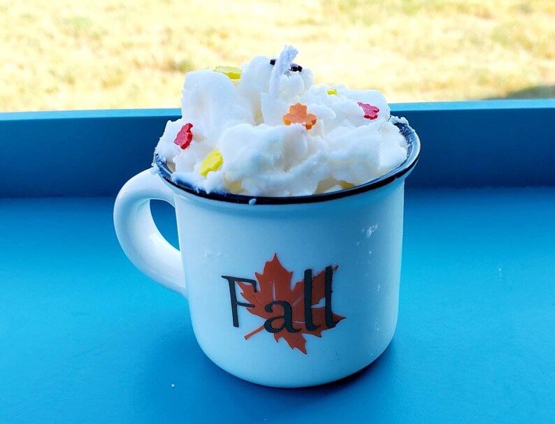 May include: A white enamel mug with a black rim and handle, filled with a white, textured candle. The mug has a "Fall" graphic with a maple leaf. The candle is decorated with colorful leaf-shaped sprinkles.
