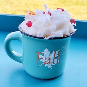 May include: A teal mug candle with a white, whipped-cream-like wax top, decorated with small, colorful leaf-shaped accents. The mug has a black rim and the word "Fall" with a leaf design on the front. The candle is set on a blue surface.
