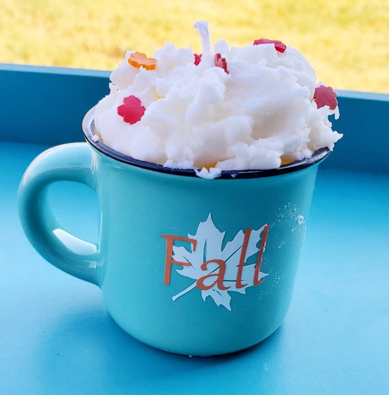 May include: A teal mug candle with a white, whipped-cream-like wax top, decorated with small, colorful leaf-shaped accents. The mug has a black rim and the word "Fall" with a leaf design on the front. The candle is set on a blue surface.