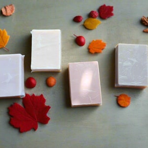 Puede incluir: Cuatro barras rectangulares de jabón en tonos blancos y rosas se disponen sobre una superficie verde apagado, junto a hojas decorativas de fieltro y pequeñas esferas rojas y naranjas. Los jabones están envueltos en plástico transparente.
