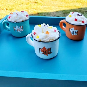 May include: Three mugs with white whipped topping and colorful sprinkles sit on a blue tray. The mugs are teal, white, and orange, each with a "Fall" leaf design. The tray is outdoors on a grassy surface.