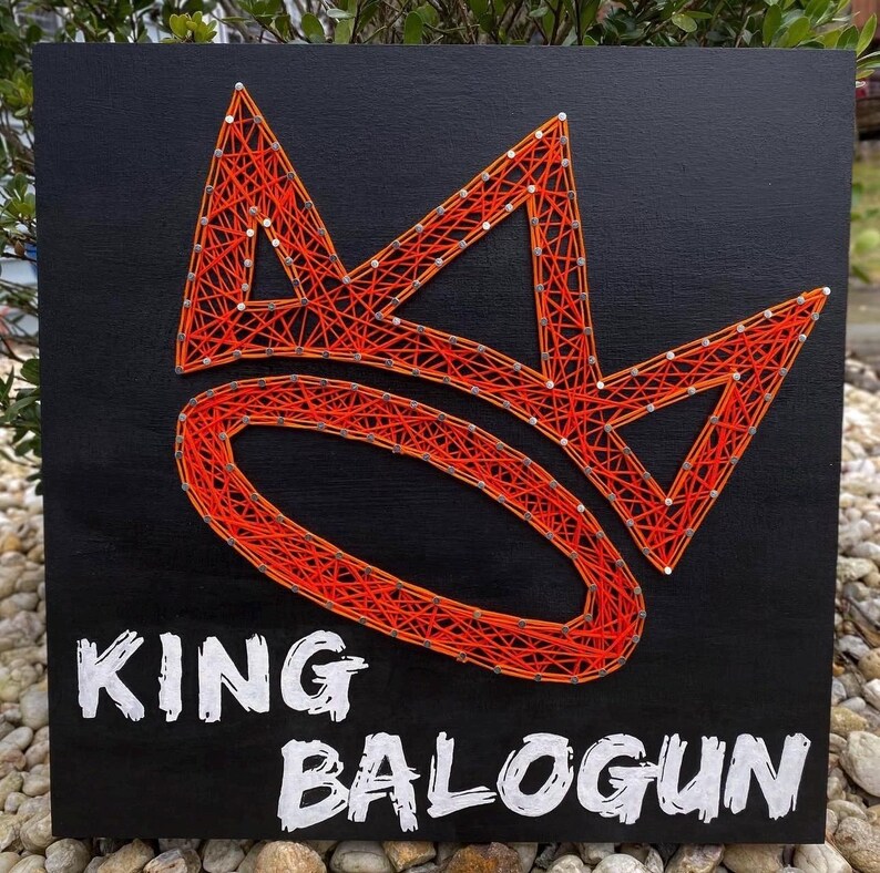 K&ouml;nnte beinhalten: Ein schwarzes Holzschild mit einem Kronen-Design aus orangefarbener Fadenkunst. Die Worte "KING BALOGUN" sind in wei&szlig;en Blockbuchstaben darunter geschrieben. Das Schild wird im Freien auf einem Bett aus kleinen Steinen ausgestellt.