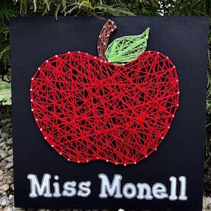 K&ouml;nnte beinhalten: Schwarzes quadratisches Schild mit einem roten String-Art-Apfel, einem gr&uuml;nen Blatt und einem braunen Stiel. Das Schild zeigt den Text "Miss Monell" in wei&szlig;er Schrift. Der Apfel ist aus rotem Faden und kleinen silbernen N&auml;geln gefertigt.