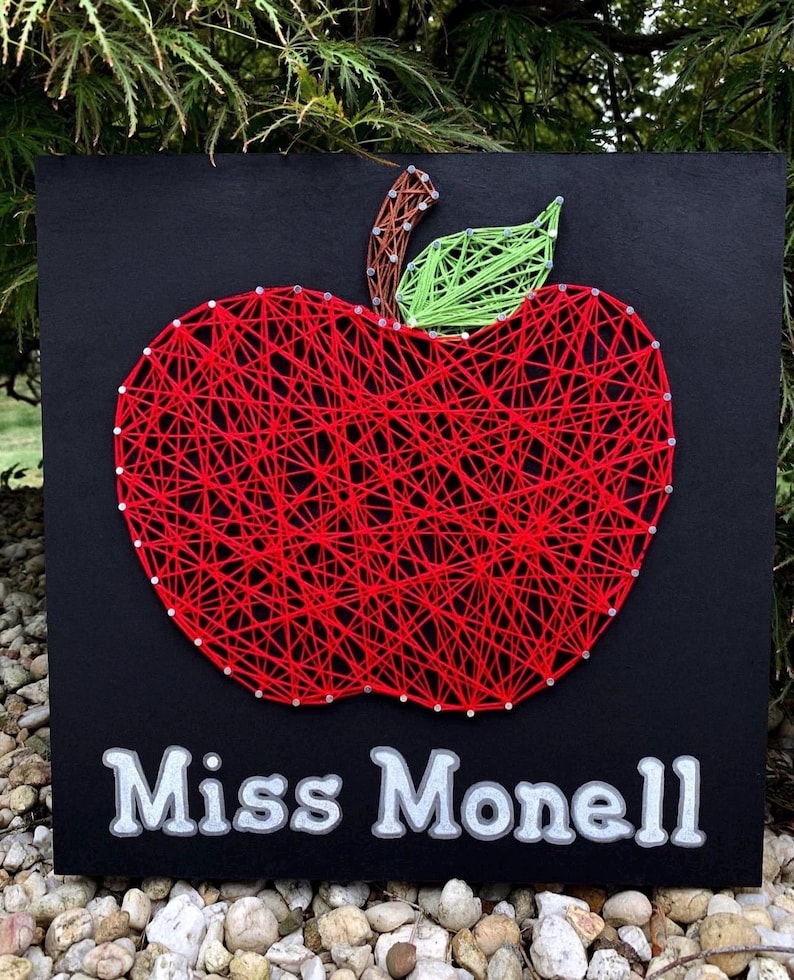 K&ouml;nnte beinhalten: Schwarzes quadratisches Schild mit einem roten String-Art-Apfel, einem gr&uuml;nen Blatt und einem braunen Stiel. Das Schild zeigt den Text "Miss Monell" in wei&szlig;er Schrift. Der Apfel ist aus rotem Faden und kleinen silbernen N&auml;geln gefertigt.
