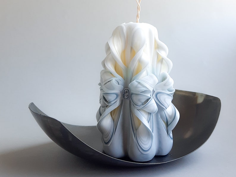 May include: A white and blue carved candle with a decorative bow. The candle is sitting on a black metal tray.