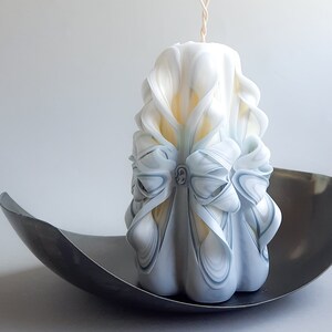 May include: A white and blue carved candle with a decorative bow. The candle is sitting on a black metal tray.