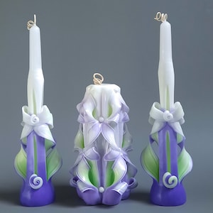 Op de afbeelding: Drie gesneden kaarsen in een wit, paars en groen kleurenschema. De kaarsen zijn versierd met bloemen en bladeren. De kaarsen zijn in een rij gerangschikt.