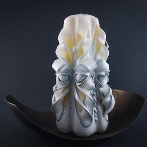 May include: A white and blue carved candle with intricate details. The candle is sitting on a dark brown metal plate.