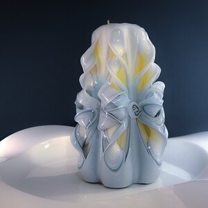 May include: A white and yellow carved candle with a decorative bow design. The candle is sitting on a white surface.