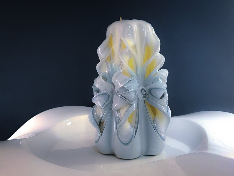 May include: A white and yellow carved candle with a decorative bow design. The candle is sitting on a white surface.