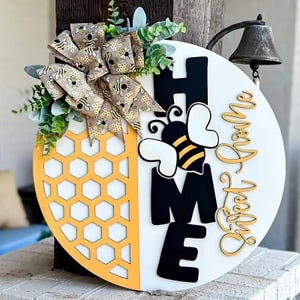 May include: A round wooden sign with a honeycomb design and the word "HOME" in black letters. A bee graphic and the words "Sweet Home" are also featured. The sign is decorated with a bow and greenery.