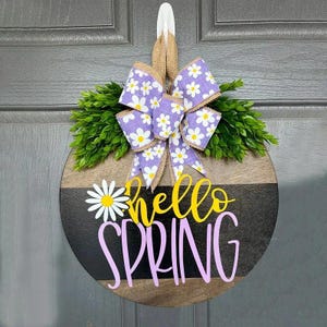 May include: Round wooden door sign with a "Hello Spring" design. The sign features a purple and white daisy-patterned bow, green foliage, and the words "hello" in yellow and "SPRING" in pink.