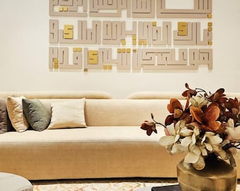 May include: A beige wall art piece featuring Arabic calligraphy in a modern design. The letters are in a light beige color with gold accents. The artwork is mounted above a beige sofa with decorative pillows. A vase of flowers sits on a table in the foreground.