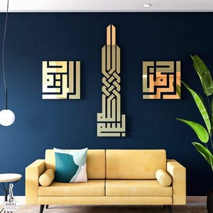 May include: Gold-colored Arabic calligraphy wall art in a modern living room setting. The artwork is displayed on a dark blue wall above a yellow sofa with a decorative pillow. A tall plant and a hanging light complete the scene.