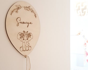 Może przedstawiać: Drewniany balon z kwiatowym wzorem i wygrawerowanym imieniem "Sumeya". Mały słoń z kwiatową koroną siedzi na balonie.