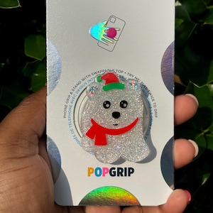 May include: A white glitter phone grip with a red scarf and a green Santa hat. The grip is a PopGrip brand and says "PopGrip" on the bottom. The background is white with black dots.