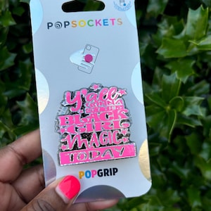 May include: A pink and black phone grip with the text "Y'all Gonna Get Hit With Black Girl Magic Today" on it. The grip is packaged on a white card with the PopSockets logo.