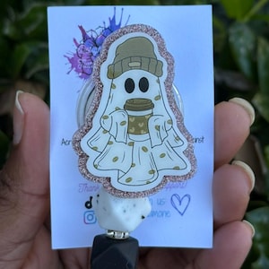 May include: A white ghost-shaped badge reel with a coffee cup and a beanie. The badge reel has a glittery border and is attached to a white and black beaded lanyard. The background is a blurred green.