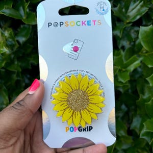 May include: A yellow sunflower PopGrip phone grip with gold glitter in the center. The PopGrip is packaged on a white card with the words "PopSockets" and "PopGrip" printed on it.