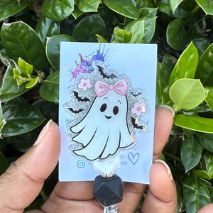 May include: A white ghost-shaped badge reel with a pink bow, flowers, and bats. The badge reel has a black and white bead design and a clear plastic clip. The background is a lush green plant.