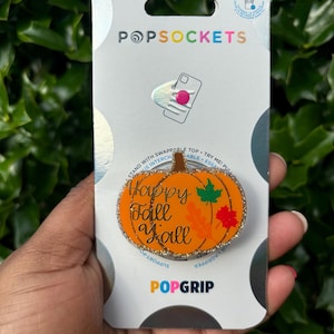 May include: A gold glitter pumpkin phone grip with the text "Happy Fall Y'all" on it. The grip is attached to a white card with the word "POPGRIP" on it.